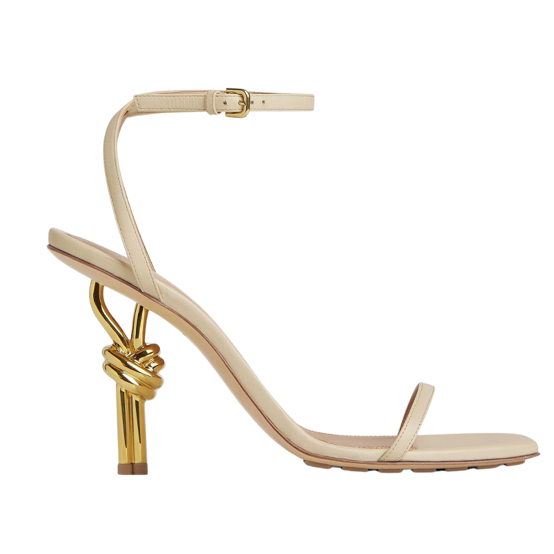 A single beige leather high-heeled sandal with a decorative gold knot detail on the heel and a matching ankle strap, shown from a side profile on a white background.