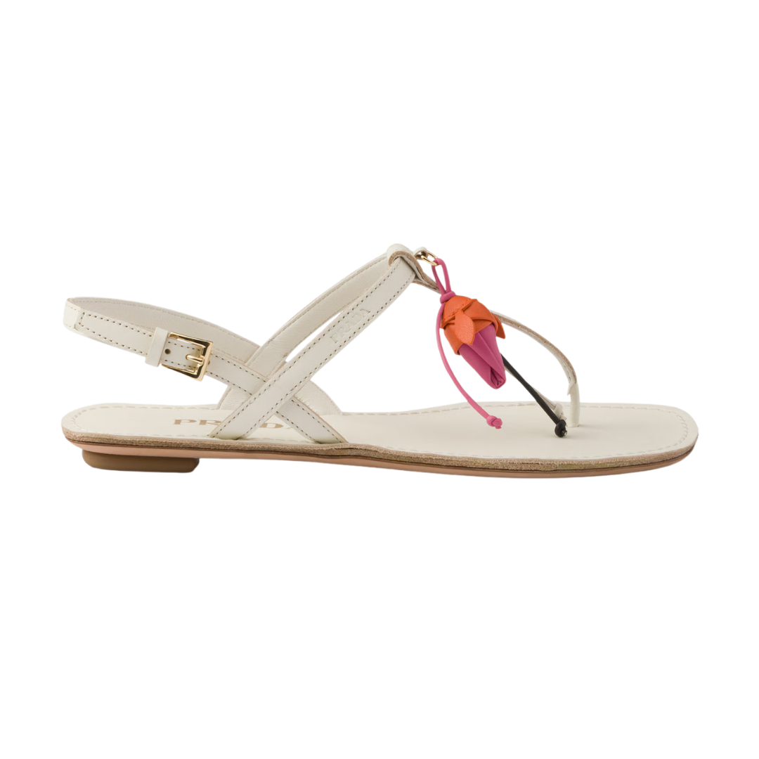 A single flat sandal in off-white leather with a buckle strap, a toe loop, and a decorative hanging charm featuring orange and pink leaf shapes, shown against a plain white background.